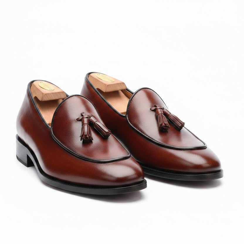 A pair of brown leather tassel loafers displayed on a dark brown surface against a black textured wall with blue lighting. A black box with gold lettering is partially visible behind the shoes.